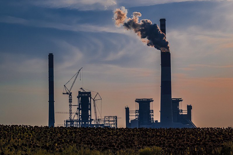 Picture of an industrial unit in a field with smoke coming out of a high funnel and into the clear sky.