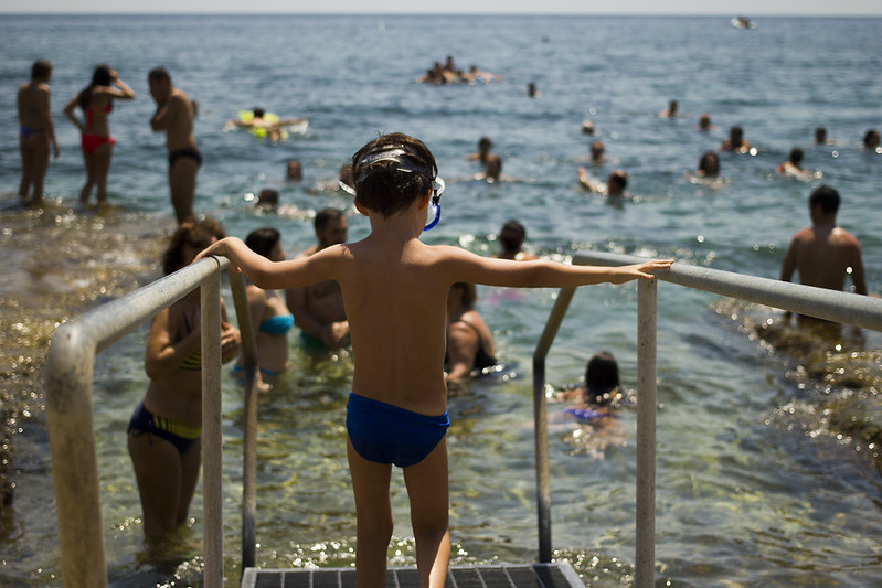 Picture of the back of a small boy in bathing suit standing on the steps leading into the sea where other people are already in the water.