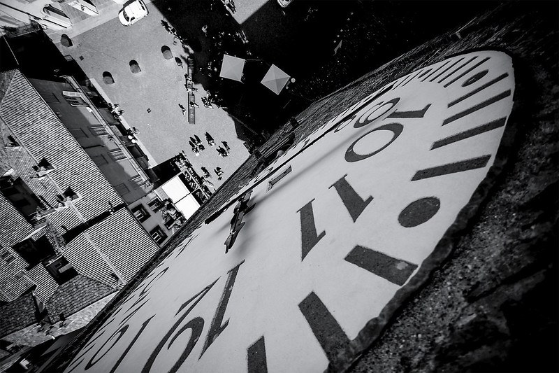 Black and white view from above a circular clock looking down into a square with a building on the left and a parked car adjacent.