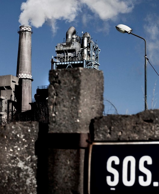 Picture from a cement wall with an industrial plant and funnel emitting smoke in the background and a sign reading SOS at the bottom right.