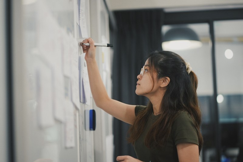 Picture of the top profile of a young brunette girl with glasses writing with a black marker on a whiteboard.