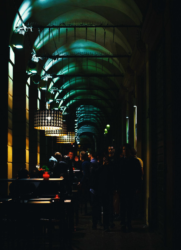 Picture of a dark arched gallery with laps visible on the left and people sitting at high tables, and a couple moving towards the camera on the left.