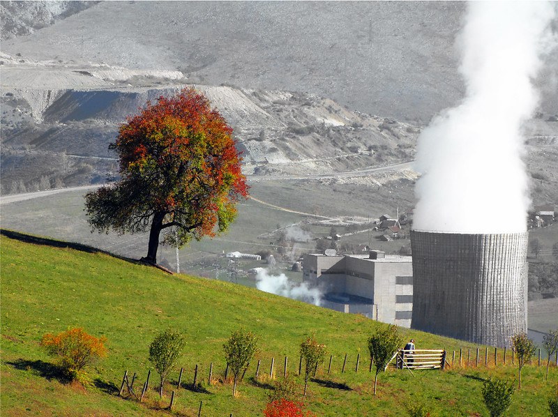 Picture of a green hillside with a large green-red tree and a man standing at a gate on the right, while a large grey smoking funnel is visible on the right in a grey hillside background.