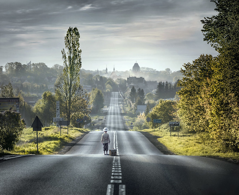 Picture of a small boy wearing a hat with a suitcase beside him standing in the middle of an up-and-down street leading to a town in the background with trees on either side of the street.